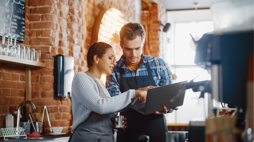 Two retail employees view important information on a laptop while working behind a barista counter